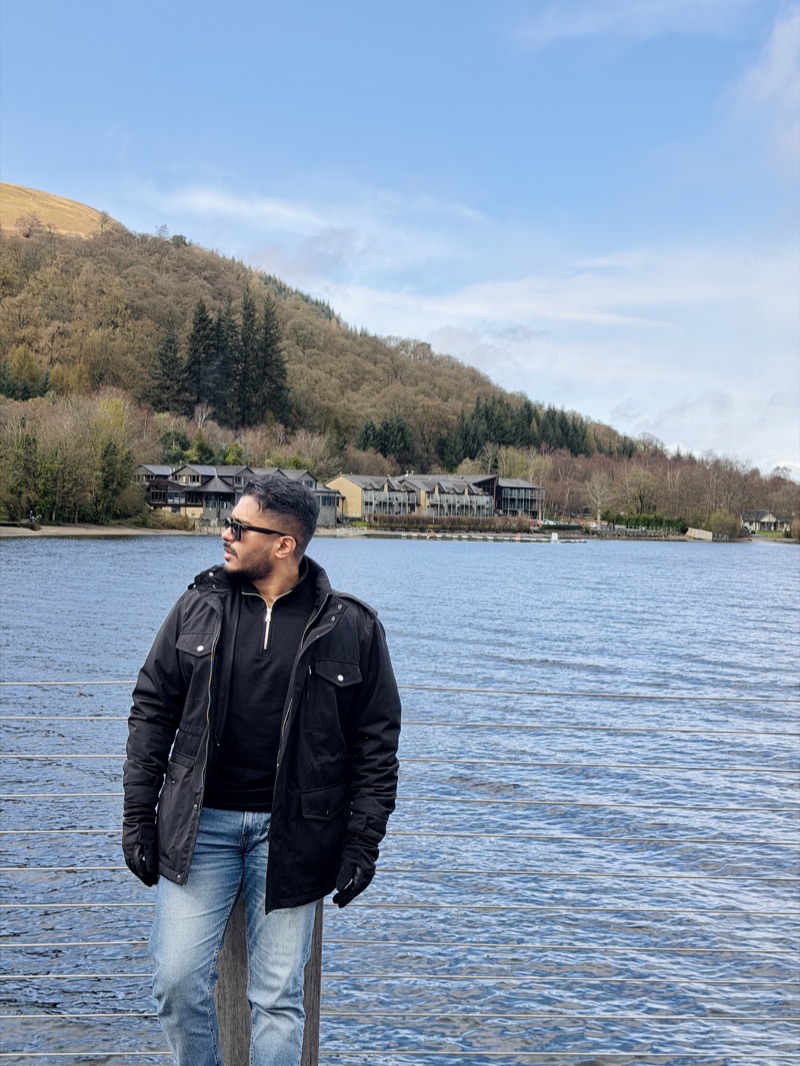 Yash Pandey standing by a loch, with forested hills and lochside lodges behind him